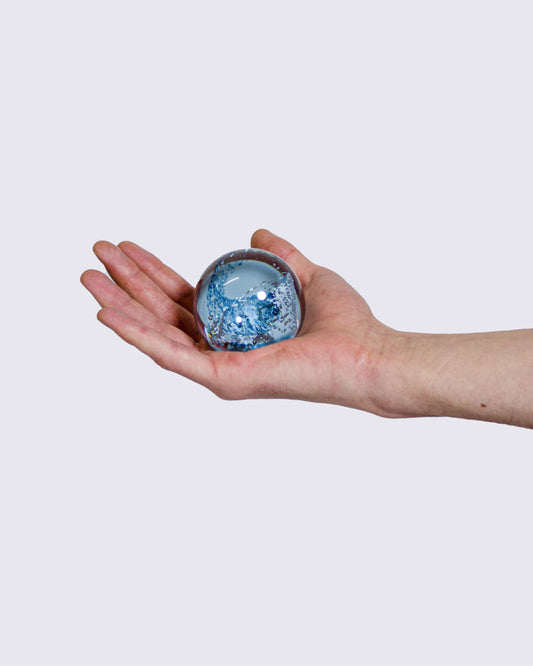 A hand holding a small, spherical glass crystal ball with blue and white elements inside, set against a solid light gray background.