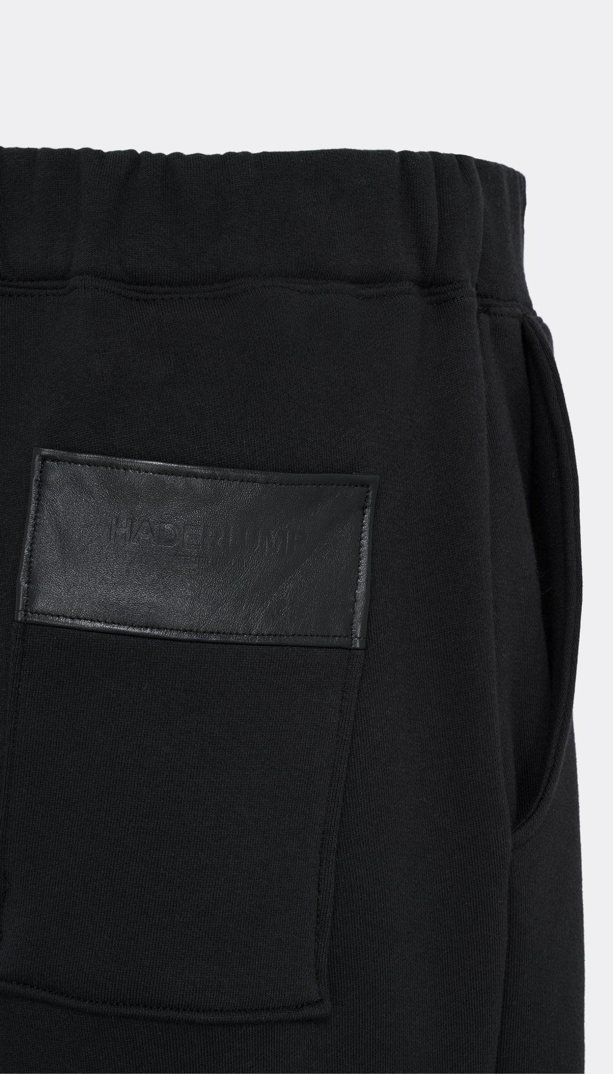 Close-up of black Haderlump pants showing a side pocket with a black leather patch that has debossed branding, set against a plain light gray background.