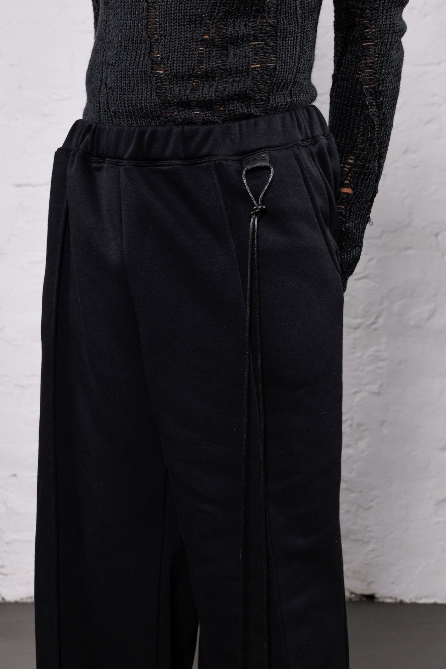 Black wide-leg trousers with a drawstring waist and side zipper detail, shown on a model wearing a dark textured top against a white brick wall.