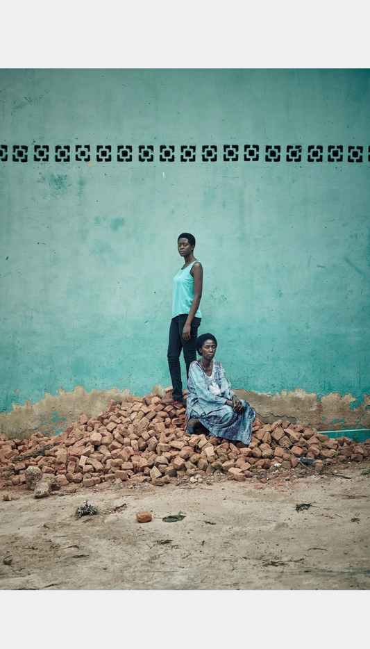 A woman in a light blue tank top and black pants stands on a pile of red bricks while a woman in a light blue patterned dress sits in front of a turquoise wall.