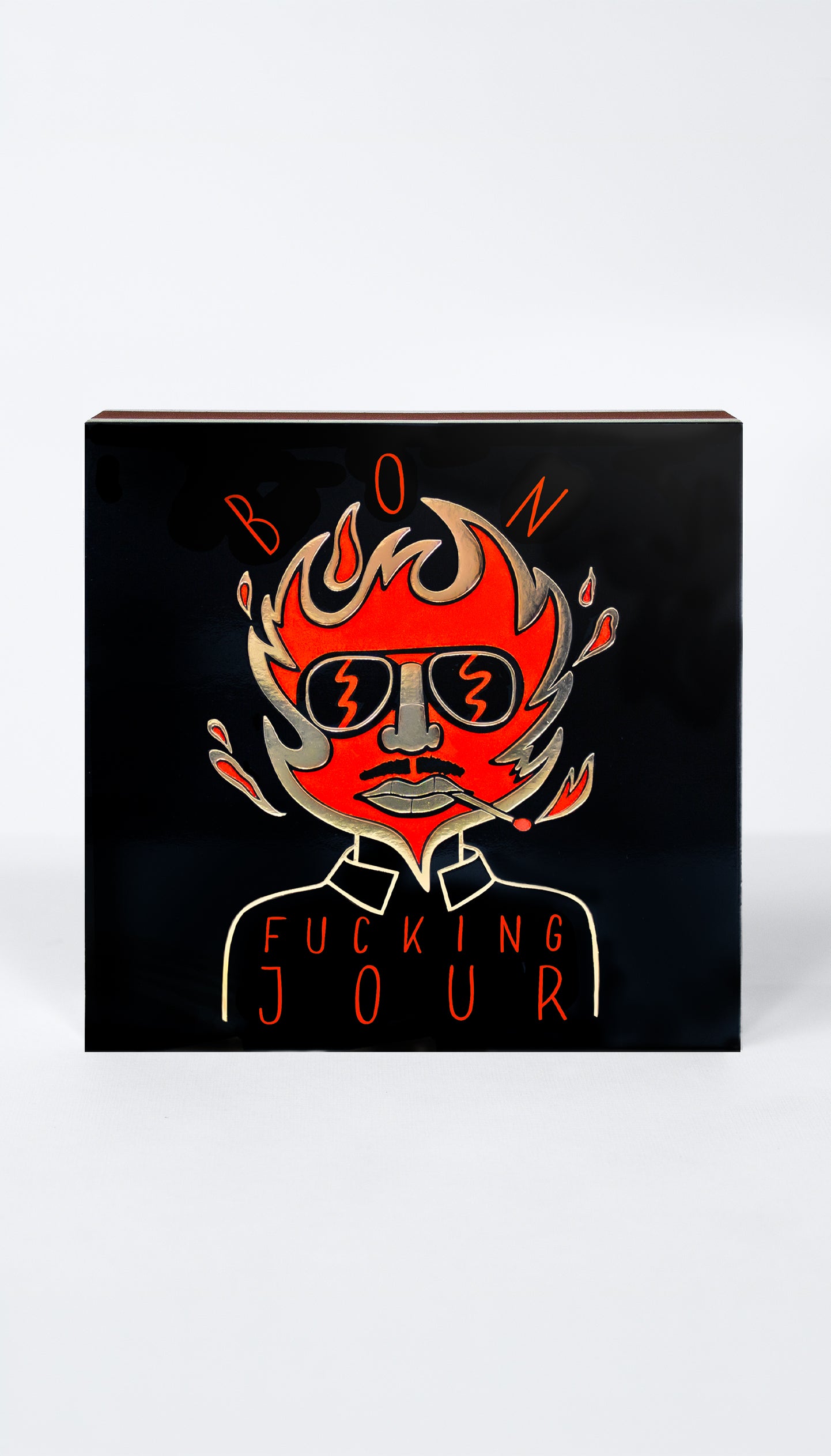 A square black matchbox featuring a stylized red and orange illustration of a face with flames and sunglasses, with the text 'BON' above and 'FUCKING JOUR' below, shown on a light gray background.