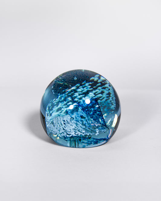 A spherical glass paperweight featuring an intricate blue and white water pattern, set against a clean light gray background.