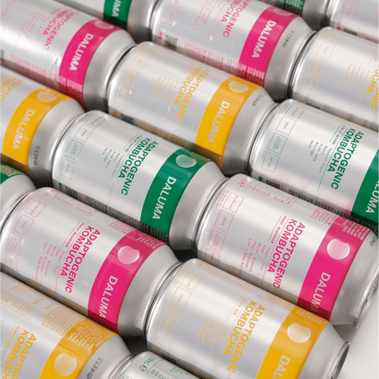 A close-up, angled view of multiple aluminum cans of Daluma Adaptogenic Kombucha arranged in a stacked pattern. The cans are metallic silver with colorful labels in pink, yellow and green.