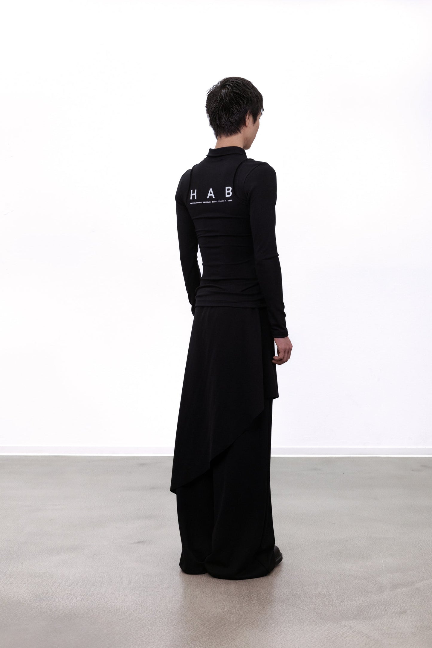 A model viewed from the back wearing a black long-sleeve mock-neck top and matching wide-leg skirt set, featuring 'HAB' printed in white on the upper back, set against a minimalist white studio background.