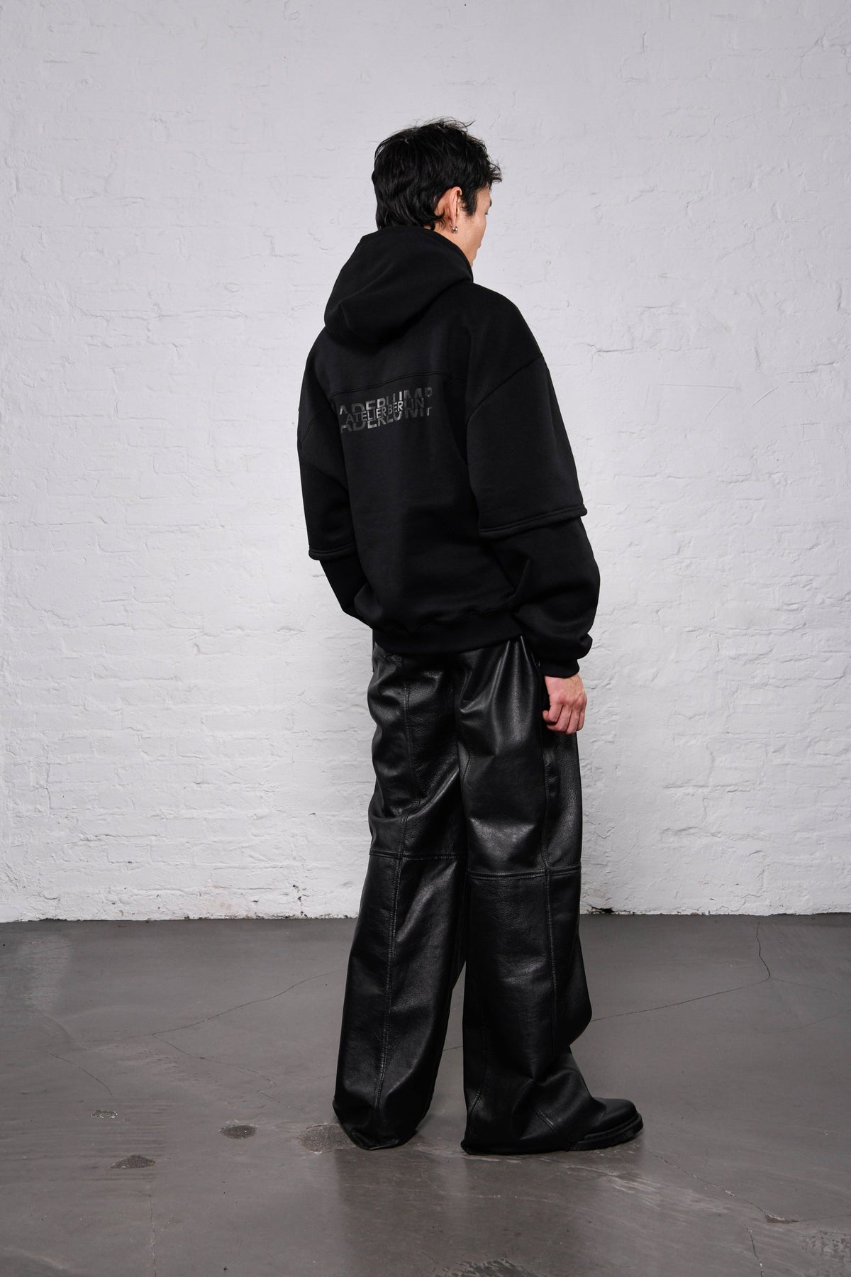 A model wearing a black oversized hoodie with a subtle graphic on the back and wide-leg black faux-leather pants, standing against a white brick wall.