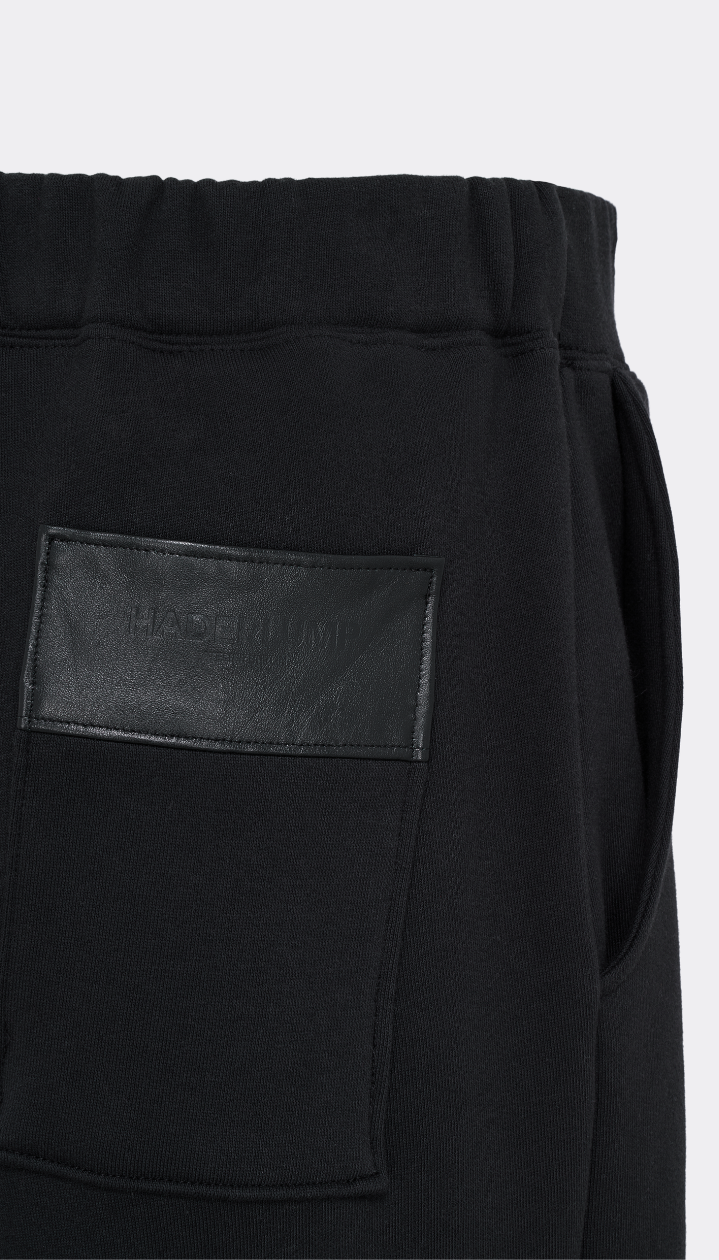 Close-up of black Haderlump pants showing a side pocket with a black leather patch that has debossed branding, set against a plain light gray background.