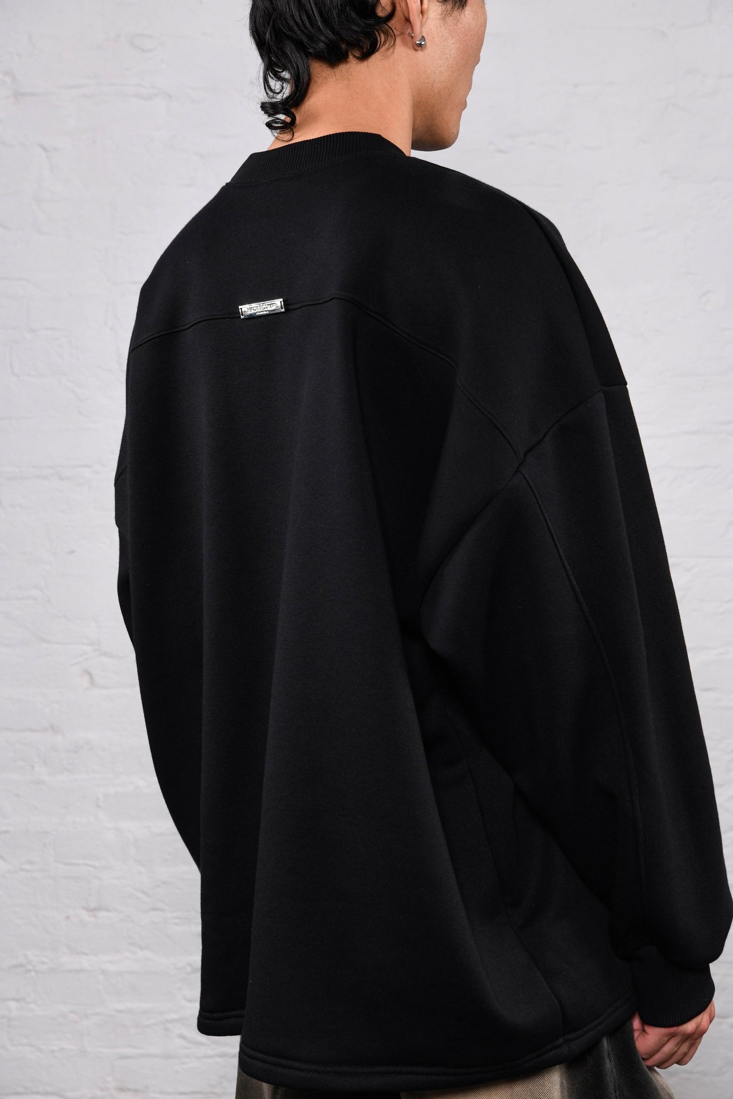 A person shown from the back wearing an oversized black crewneck sweatshirt with a small white rectangular label on the upper back, set against a white brick wall.