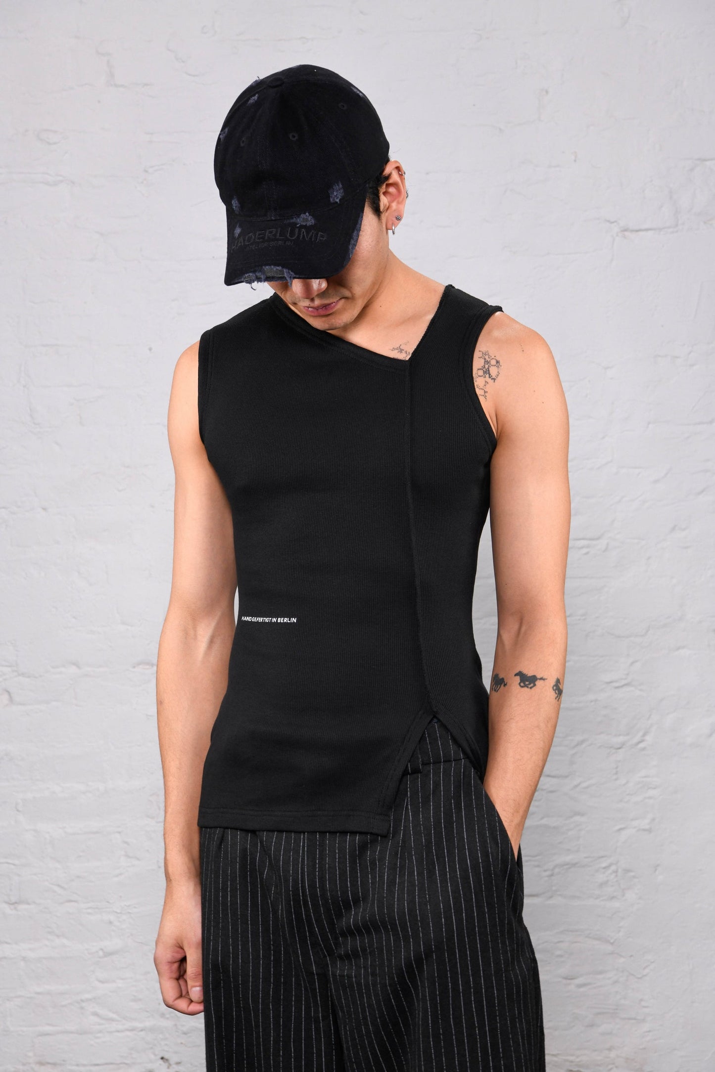 A man wearing a black asymmetrical sleeveless knit top, black pinstripe trousers, and a black baseball cap, standing against a white brick wall.