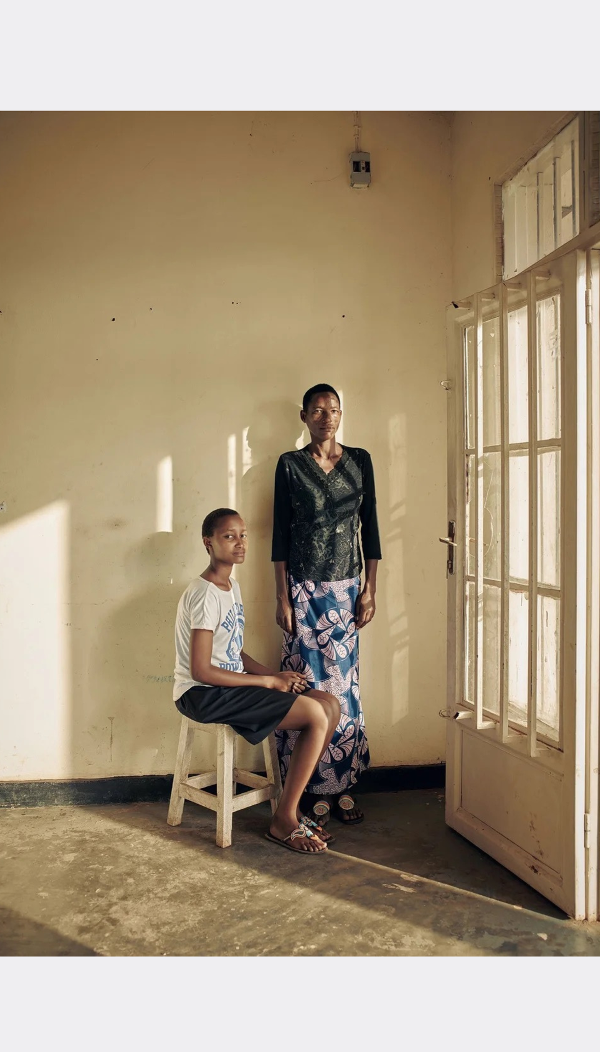 A woman in a white t-shirt and dark shorts sits on a small white stool, while a woman in a black top and blue patterned skirt stands nearby in a sunlit room with beige walls and a wooden door.