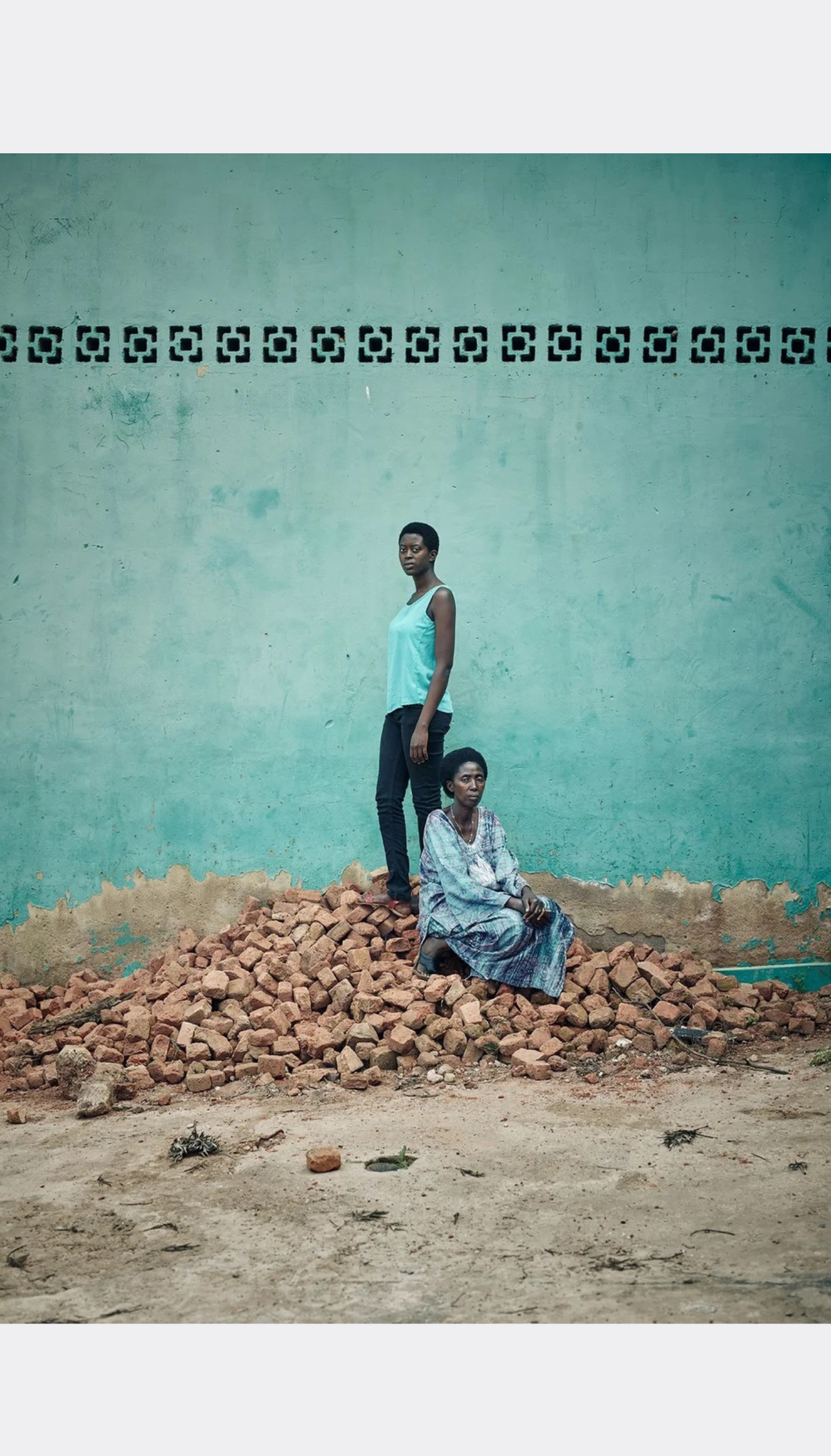 A woman in a light blue tank top and black pants stands on a pile of red bricks while a woman in a light blue patterned dress sits in front of a turquoise wall.