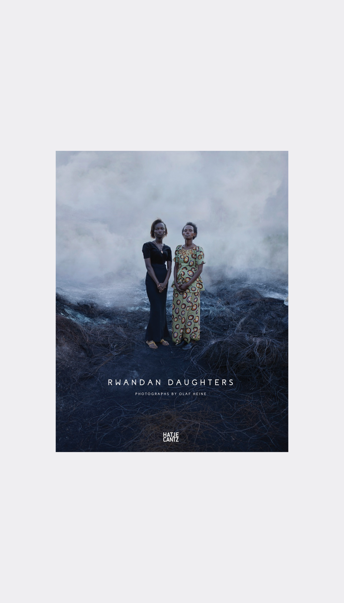 A book or album cover titled 'RWANDAN DAUGHTERS' with a moody photograph of two women standing on a dark, misty landscape. The cover is centered on a light gray background by Olaf Heine