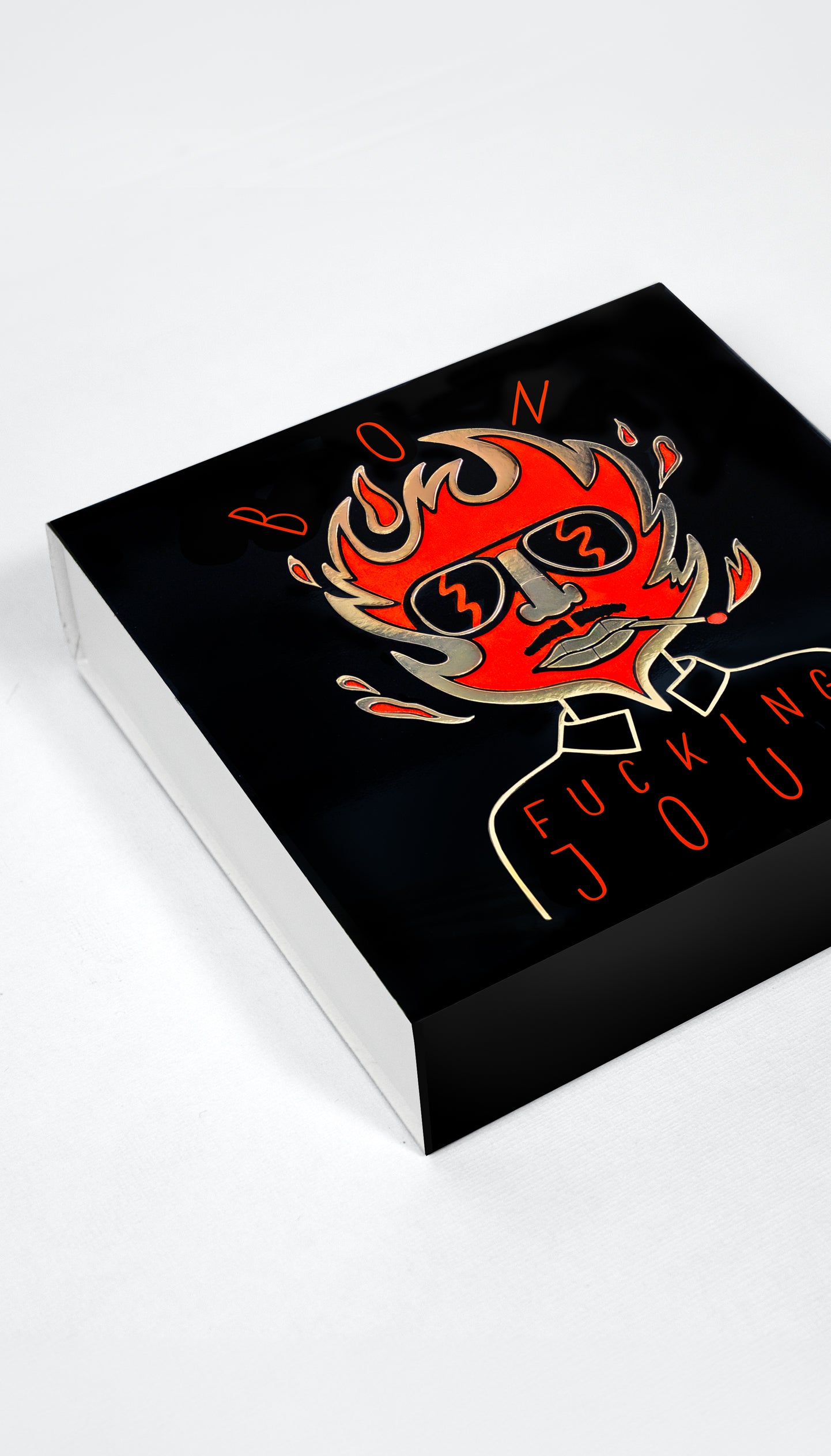 A black rectangular matchbox or shown at an angle on a white background, featuring a stylized red and orange flaming face graphic with the words 'BON' and 'FUCKING JOUR' in red and orange lettering.