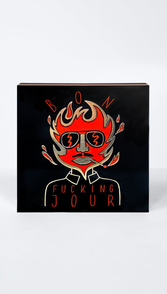 A square black matchbox featuring a stylized red and orange illustration of a face with flames and sunglasses, with the text 'BON' above and 'FUCKING JOUR' below, shown on a light gray background.