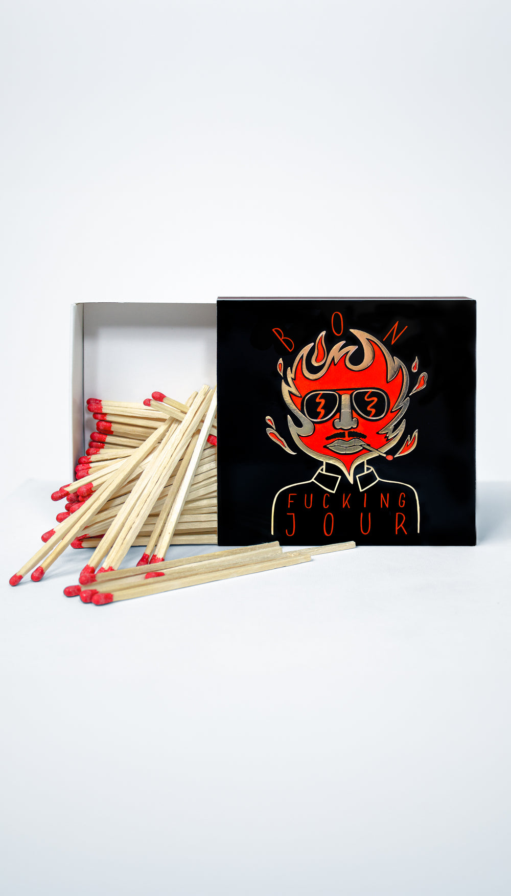A set of wooden matches with red tips arranged in front of a black matchbox lid featuring a stylized red and orange flame-faced graphic with the text 'BON FUCKING JOUR'.