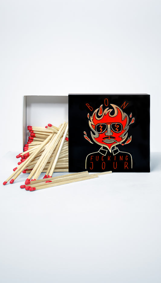 A set of wooden matches with red tips arranged in front of a black matchbox lid featuring a stylized red and orange flame-faced graphic with the text 'BON FUCKING JOUR'.