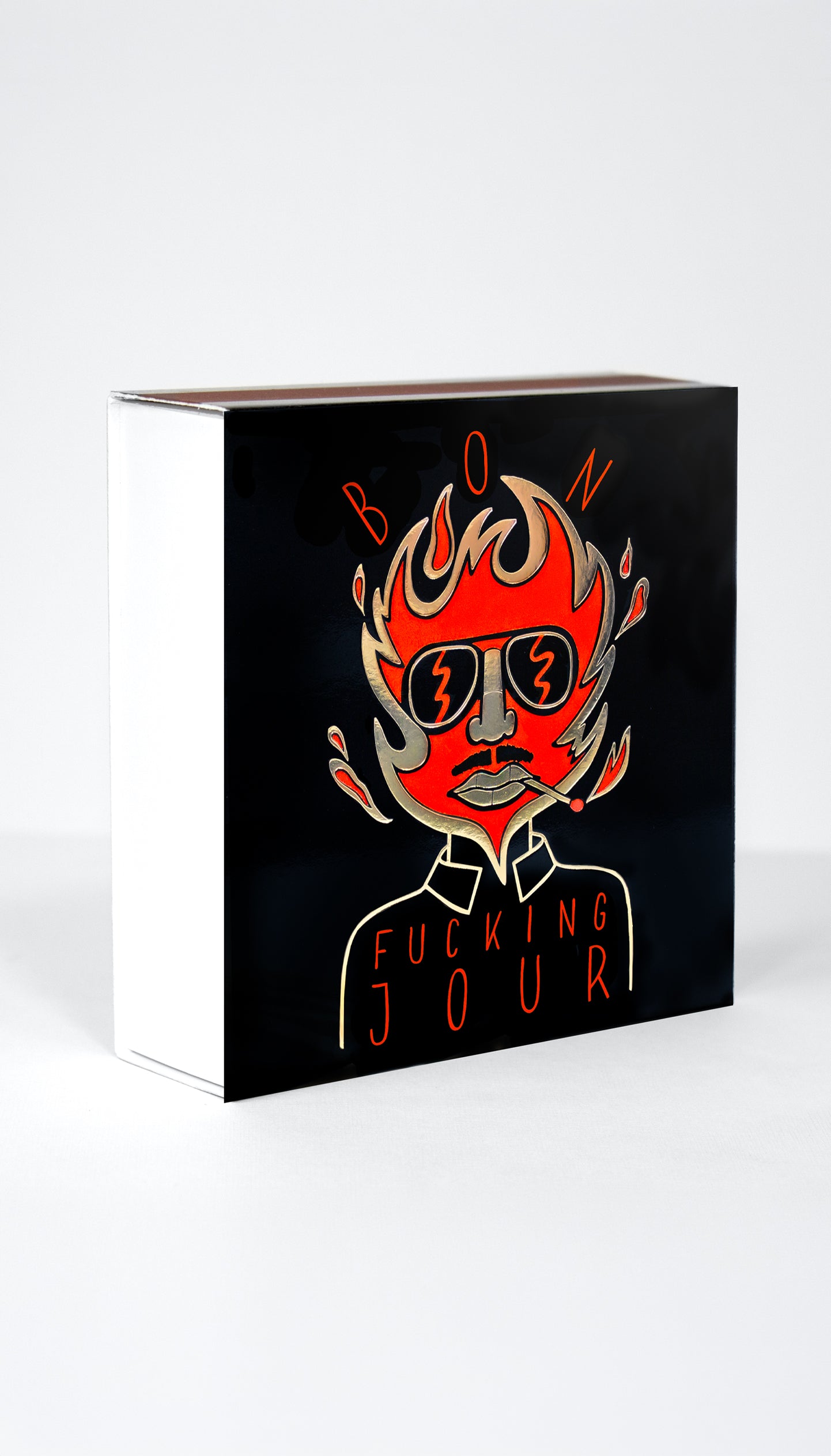 A black rectangular matchbox with a stylized illustration of a face in sunglasses with flames rising from the head, and the text 'BON FUCKING JOUR' in red and orange tones, shown on a light gray background.